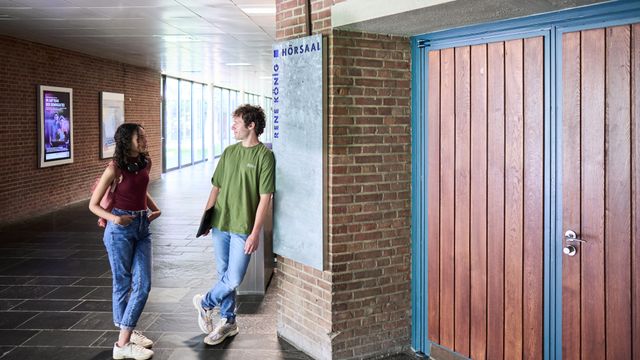 Studierende unterhalten sich im Flur vor einem Hörsaal auf dem Campus.