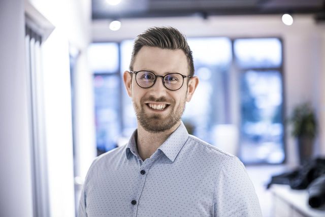 Porträt eines lächelnden Mannes mit Brille im Business-Casual-Outfit in einem modernen Büro.