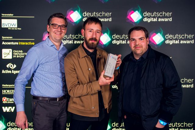 Benjamin Arndt, Jan Teunissen und Marcel Schindler präsentieren stolz ihren gewonnenen Award vor dem Hintergrund der Deutschen Digital Awards.