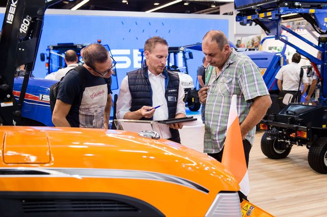 Gesprächssituation zwischen einem Produktmanager und zwei Kunden auf einem Messestand mit Traktoren