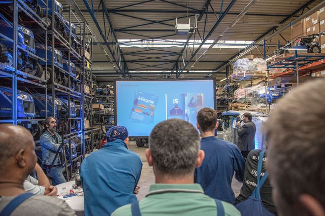 Menschenmenge steht in einer Lagerhalle mit Traktoren und schaut auf eine Leinwand mit einer Präsentation, die Broschüren im neuen Corporate Design zeigt 