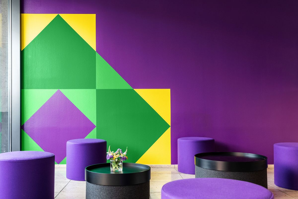 Die Sitzmöbel und Tische im Foyer greifen die geometrische Formsprache und die Farben passend zur Wandgestaltung mit dem Keyvisual auf. Die neue Wandgestaltung im Foyer des „Hauses der Kirche“ mit Keyvisual
