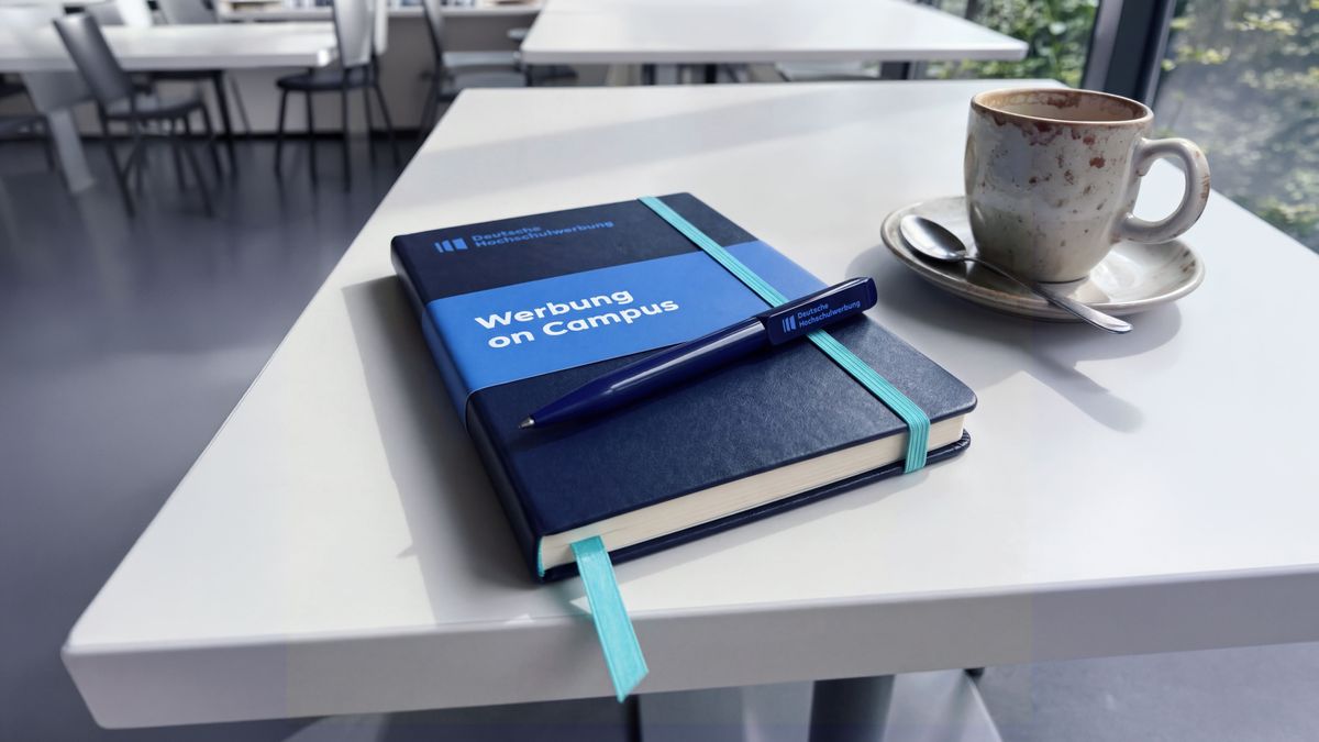 Gebrandetes Notizbuch und Kugelschreiber der Deutschen Hochschulwerbung auf einem Tisch.