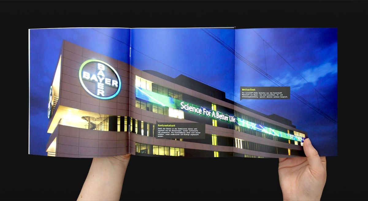 Aufgeklappte Broschüre mit einer LED-Wand am Gebäude von Bayer Unternehmen