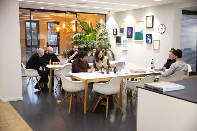 In einer offenen Büroküche sitzen mehrere Personen verteilt an Tischen und sind in Gespräche vertieft